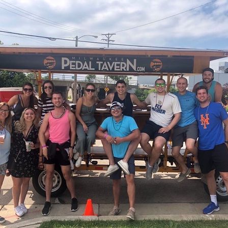 Group of friends on a Milwaukee pedal pub (party pedal bar) on a sunny day, smiling and holding drinks in casual summer clothes.