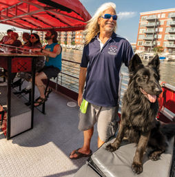 Smiling man with long hair and sunglasses stands with a large black dog on a sunny Milwaukee riverboat tour, passengers at a bar and waterfront apartments in the background.