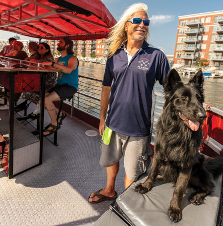 Smiling man with long hair and sunglasses stands with a large black dog on a sunny Milwaukee riverboat tour, passengers at a bar and waterfront apartments in the background.