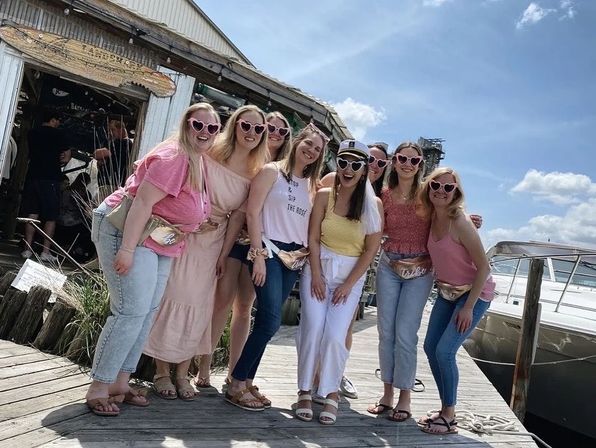 Group of eight women in heart-shaped sunglasses and summer outfits posing on a wooden marina dock beside a boat under a sunny blue sky.