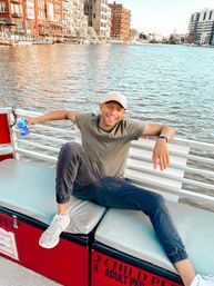 Smiling person in a baseball cap lounging on a riverboat bench, holding a blue beer can with city waterfront buildings across calm water in the background.