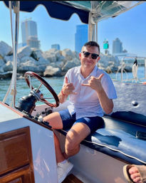 Smiling person in sunglasses and casual shirt sitting at a small boat helm, making a shaka gesture with a rocky breakwater and coastal city skyline under a sunny sky in the background.