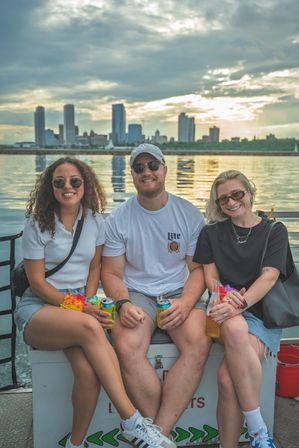 Three friends smiling on a sunset boat ride, holding drinks with a reflective downtown skyline and calm water in the background.