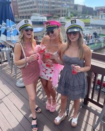 Three friends in captain hats and sunglasses toast with pink cocktails on a sunny waterfront deck overlooking boats and city buildings.
