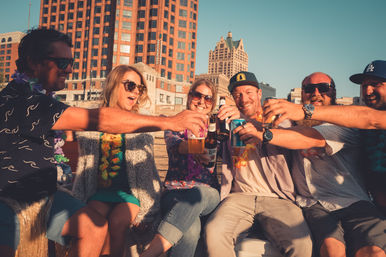 Six friends wearing leis toast with beers and cans on a sunny downtown waterfront, smiling against a city skyline.