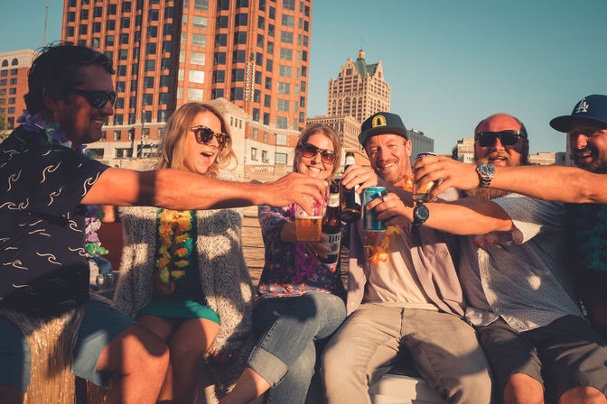 Six friends wearing leis toast with beers and cans on a sunny downtown waterfront, smiling against a city skyline.