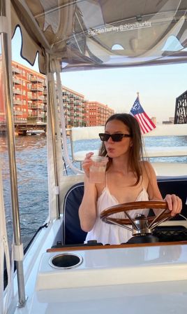 Person in sunglasses and white sundress steering a small motorboat and sipping a drink, American flag at the stern with red-brick waterfront apartments and a bridge at golden hour.
