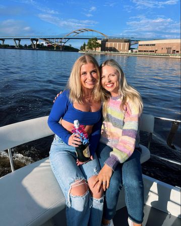 Two friends smiling on a boat at a sunny riverfront, holding a ribbon-decorated champagne bottle with an arched bridge and waterfront buildings in the background.