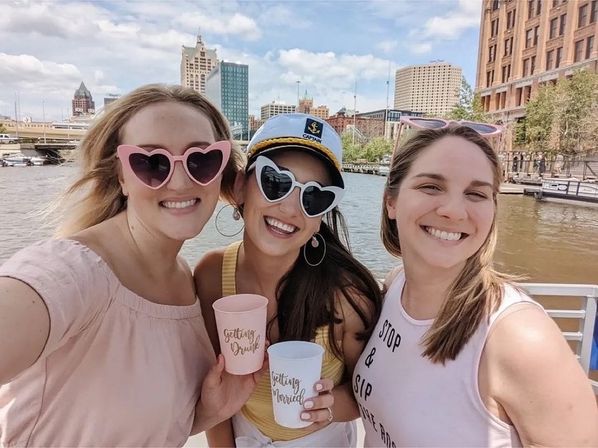 Three smiling women on a sunny downtown riverboat cruise, wearing heart-shaped sunglasses and a captain hat, holding pastel celebratory cups with a city skyline behind them.