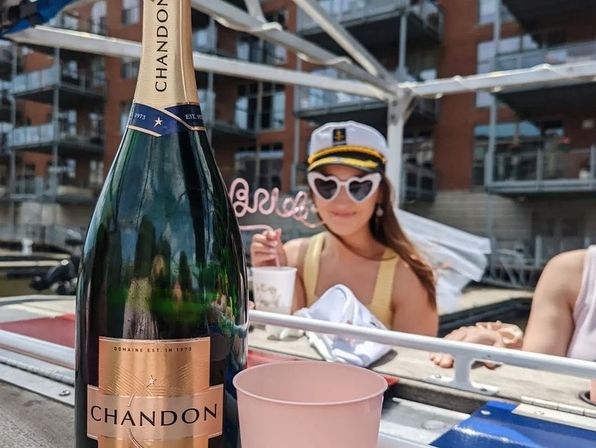 Sunny dockside boat party: large champagne bottle and pink cup on a table, person in a captain's hat and heart-shaped sunglasses in front of urban marina apartments