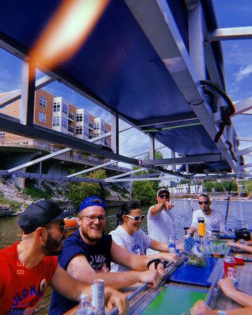 Group of friends laughing and sipping drinks at a covered party boat bar, cruising an urban waterfront past riverside apartments and a bridge on a sunny summer day.