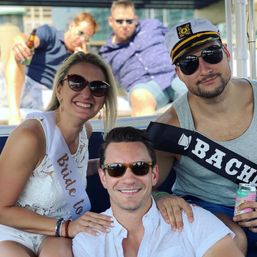 Smiling friends on a sunny boat: bride-to-be in white lace dress and sash, man in captain’s hat with a 'Bachelor' sash, sunglasses and canned drinks — casual summer bachelorette boat party