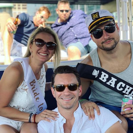 Smiling friends on a sunny boat: bride-to-be in white lace dress and sash, man in captain’s hat with a 'Bachelor' sash, sunglasses and canned drinks — casual summer bachelorette boat party