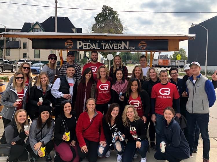 Group of about 25 adults smiling and posing around a pedal-powered party bar in Milwaukee, many wearing matching red shirts and holding drinks on a city street.