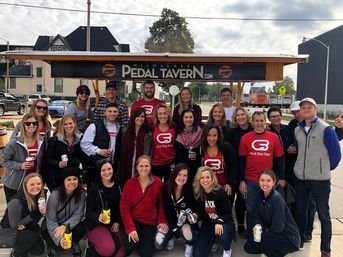 Group of about 25 adults smiling and posing around a pedal-powered party bar in Milwaukee, many wearing matching red shirts and holding drinks on a city street.