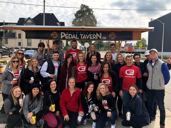 Group of about 25 adults smiling and posing around a pedal-powered party bar in Milwaukee, many wearing matching red shirts and holding drinks on a city street.