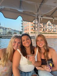 Three smiling friends on a boat at a sunny city waterfront, holding cocktails beneath a canopy with modern waterfront apartments in the background.