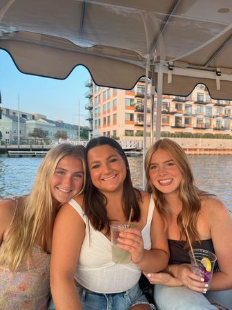 Three smiling friends on a boat at a sunny city waterfront, holding cocktails beneath a canopy with modern waterfront apartments in the background.