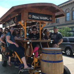Group of friends laughing and pedaling a wooden pedal-powered bar bike on a sunny downtown Milwaukee street, seated around a central bar with drinks and a large wooden barrel