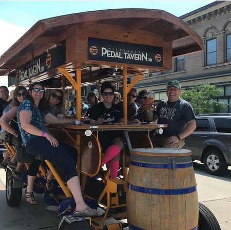 Group of friends laughing and pedaling a wooden pedal-powered bar bike on a sunny downtown Milwaukee street, seated around a central bar with drinks and a large wooden barrel