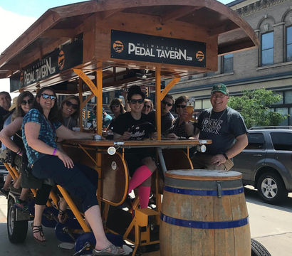 Group of friends laughing and pedaling a wooden pedal-powered bar bike on a sunny downtown Milwaukee street, seated around a central bar with drinks and a large wooden barrel