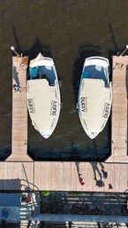 Aerial drone view of two white boats with matching beige canvas covers docked between wooden marina piers on brown river water; people and long shadows stretch across the sunlit dock.