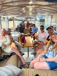 Smiling group of friends on a covered pontoon boat at a sunny marina, gathered around a wooden table with drinks — relaxed summer waterfront boat party scene.