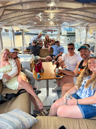 Smiling group of friends on a covered pontoon boat at a sunny marina, gathered around a wooden table with drinks — relaxed summer waterfront boat party scene.