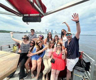 Smiling group of friends posing and waving on an open-deck boat tour with a distant city skyline and calm harbor water on a sunny day
