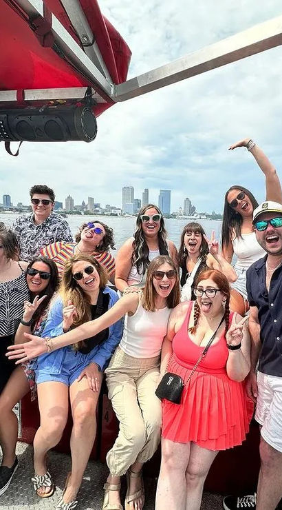 Smiling group of friends posing and waving on an open-deck boat tour with a distant city skyline and calm harbor water on a sunny day