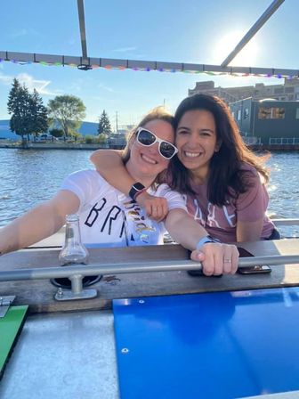 Two friends smiling and hugging on a sunny waterfront boat ride, one wearing white sunglasses and a bride sash, marina buildings and trees in the background.