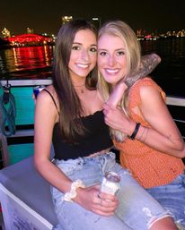 Two smiling friends embracing on a night boat ride, holding canned drinks, wearing casual summer outfits with a visible arm tattoo, waterfront city skyline and red-lit building reflecting on the water