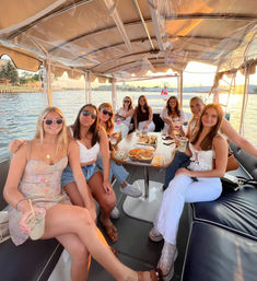 Smiling friends on a covered pontoon boat at sunset, enjoying drinks and snacks with waterfront city skyline and American flag in the background