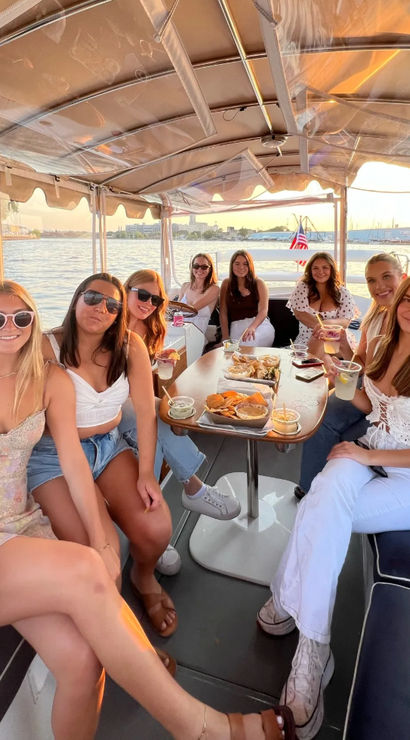 Smiling friends on a covered pontoon boat at sunset, enjoying drinks and snacks with waterfront city skyline and American flag in the background