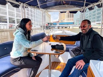 Two people toasting with beer on a covered boat during a downtown river cruise, city buildings and a bridge lining the urban waterfront