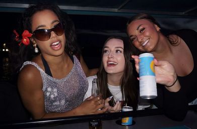 Three friends laughing on a nighttime boat deck party by the water — one wearing sunglasses and a red flower in her hair with a sparkly top, the center woman holding a bottle and smiling up, and the right woman leaning in offering a canned drink to the camera.