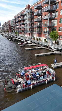 Red paddlewheel party boat with passengers cruising a brown river past wooden docks and a long row of red-brick waterfront apartments with balconies on a sunny urban riverfront
