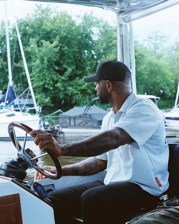 Person in a baseball cap with tattooed arms steering a motorboat at a marina, sailboats and green shoreline visible in the background.