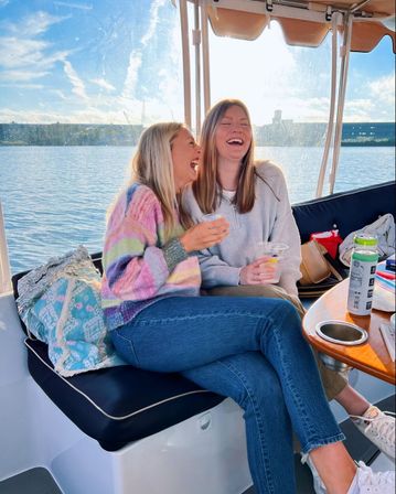 Two friends laughing on a sunlit boat near a marina, sipping drinks — one in a colorful striped sweater and jeans, waterfront skyline in the background.