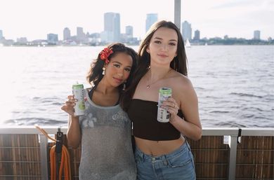 Two friends on a sunny boat deck by the waterfront, holding canned drinks with a city skyline and sailboats on the water behind them.