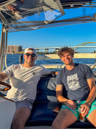 Two friends smiling on a motorboat under a clear blue sky, waterfront city and yellow-arched bridge in the background, casual summer clothes with a canned drink — sunny boating day.
