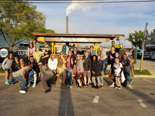 Large group of smiling adults posing around a yellow pedal-powered party bike on a sunny city street, with a tall industrial smokestack emitting white smoke and trees in the background; late afternoon light and long shadows.