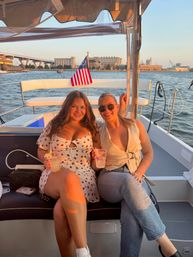 Two friends smiling on a sunset harbor boat cruise, holding cocktails on a small leisure boat with an American flag and waterfront skyline in the background.