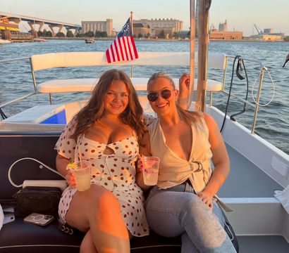 Two friends smiling on a sunset harbor boat cruise, holding cocktails on a small leisure boat with an American flag and waterfront skyline in the background.