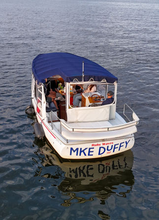 Small covered pontoon boat with a navy canopy, people chatting and snacking around a table while the captain steers, floating on calm water with a clear reflection.