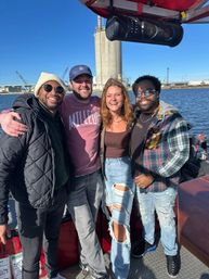 Four friends smiling on a sunny boat ride along an industrial waterfront harbor with blue sky, casual jackets and ripped jeans.