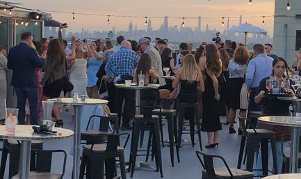 Rooftop cocktail party at sunset with guests mingling around high tables and stools under string lights, city skyline silhouette in the background