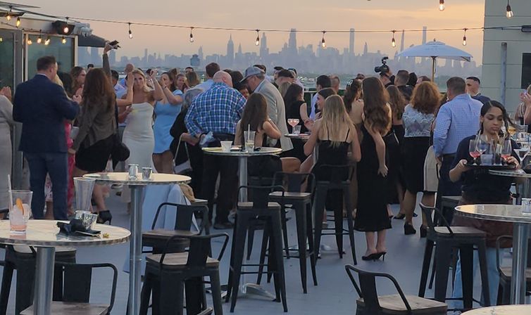 Rooftop cocktail party at sunset with guests mingling around high tables and stools under string lights, city skyline silhouette in the background