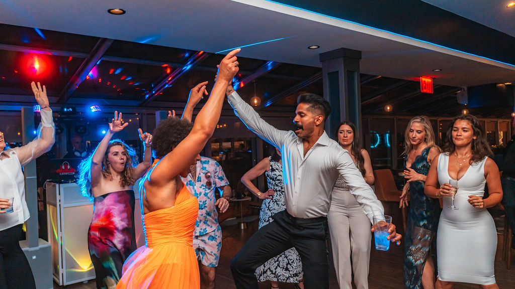 Group of adults dancing at an indoor party under colorful disco lights — a man in a white shirt leads a woman in a bright orange dress while guests in cocktail attire cheer and hold drinks.