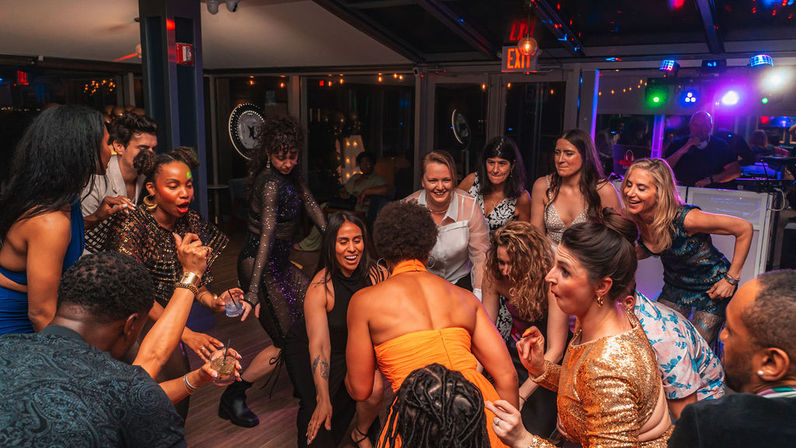 Energetic group of adults in festive attire dancing and cheering around a central dancer on an indoor party dance floor with colorful disco lights and a DJ booth in the background